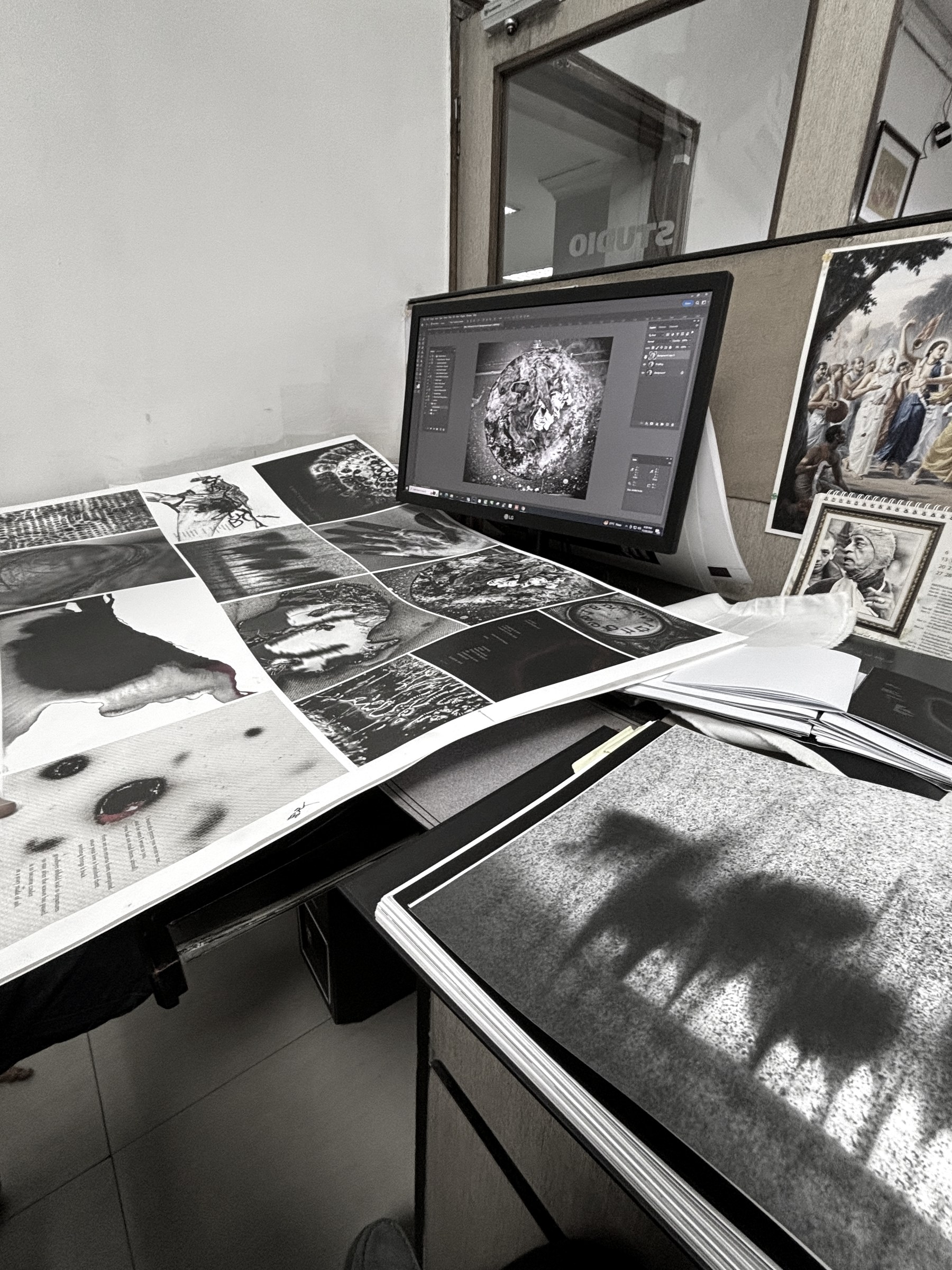 Proof sheets spread across a studio desk beside a monitor, the printer's workshop beyond