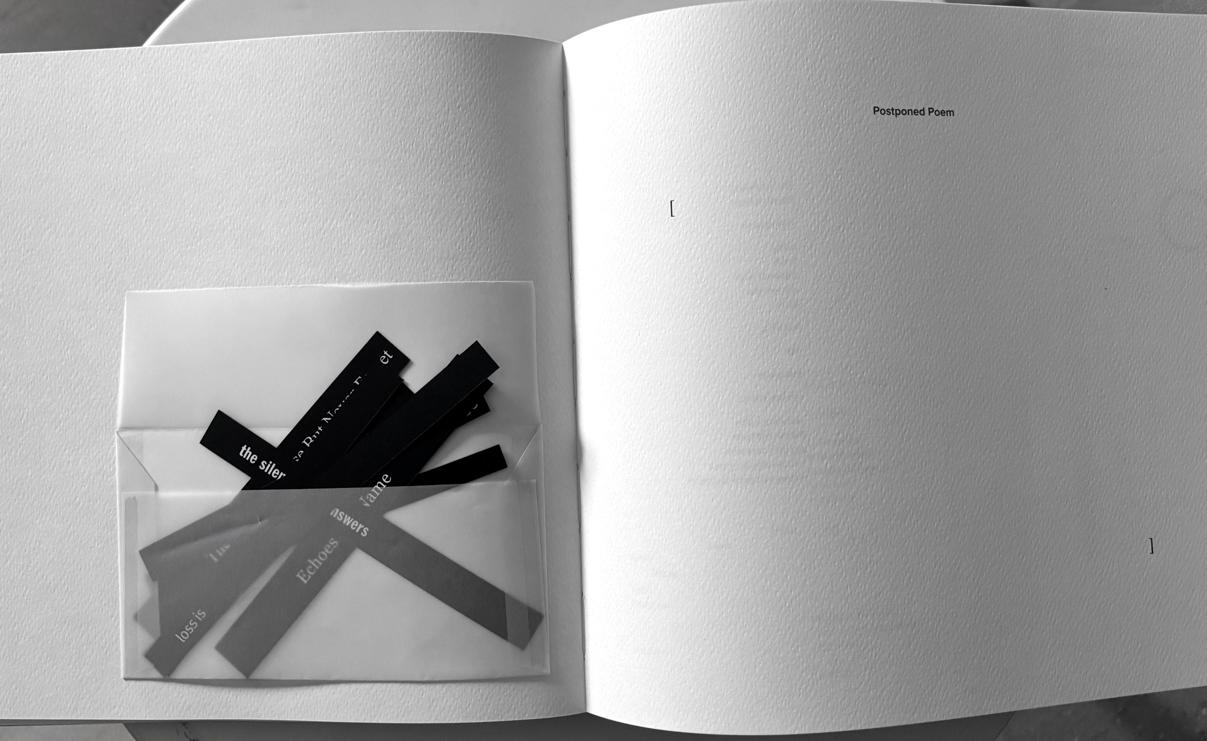 A spread with a glassine pocket holding loose printed strips of text, facing an empty page marked only with two brackets