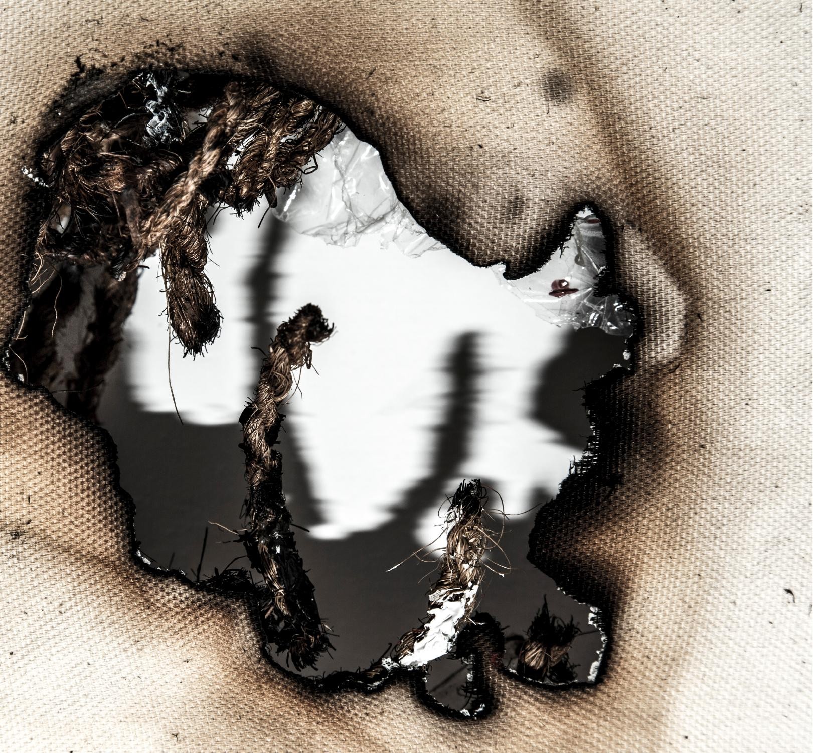 A burned aperture in cloth revealing rope and metallic paper beneath